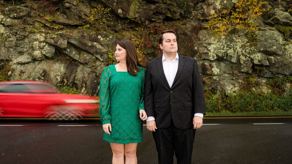 Couple standing stoically looking in opposite directions in the Shenandoah National Park as a car blurs past behind them on the Skyline Drive