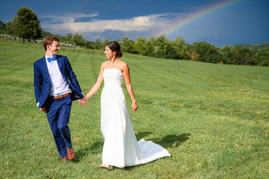 Couple walking in a field in front of a beautiful rainbow at their Veritas Vineyard wedding photography at the winery